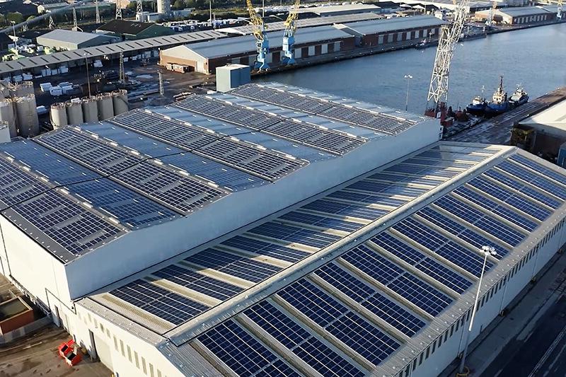 A view of a warehouse roof from a high elevation, with blue solar panels covering the roof. The warehouse is in an industrial area next to a river